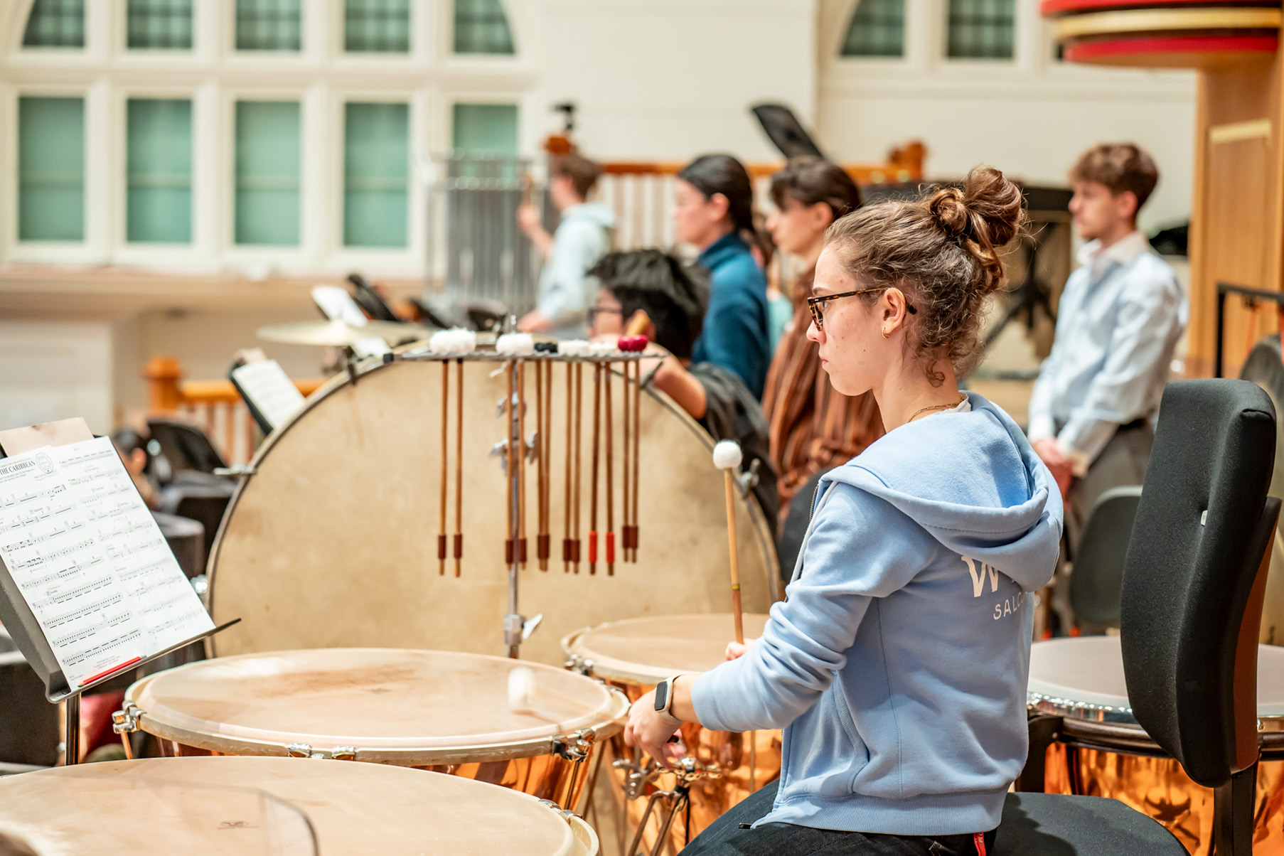 A female student playing on a timpani in an orchestra rehearsal.